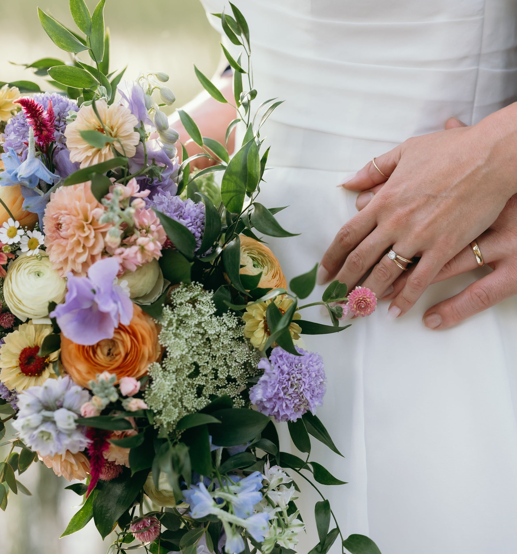 Close-up of a bride in a white wedding dress holding a colorful floral bouquet, with a groom in a navy suit resting his hands on her waist. The bride is wearing two gold wedding bands.