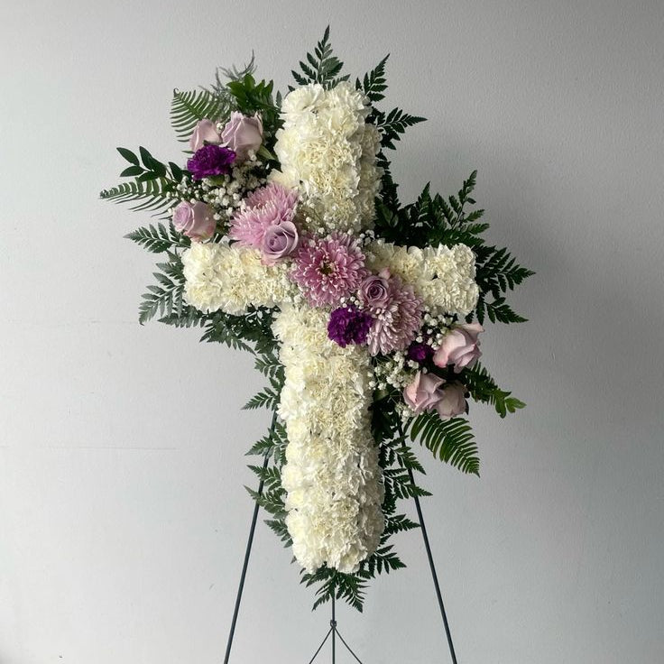 Floral cross arrangement with white and purple flowers on a stand against a plain background. An example of funeral flowers.