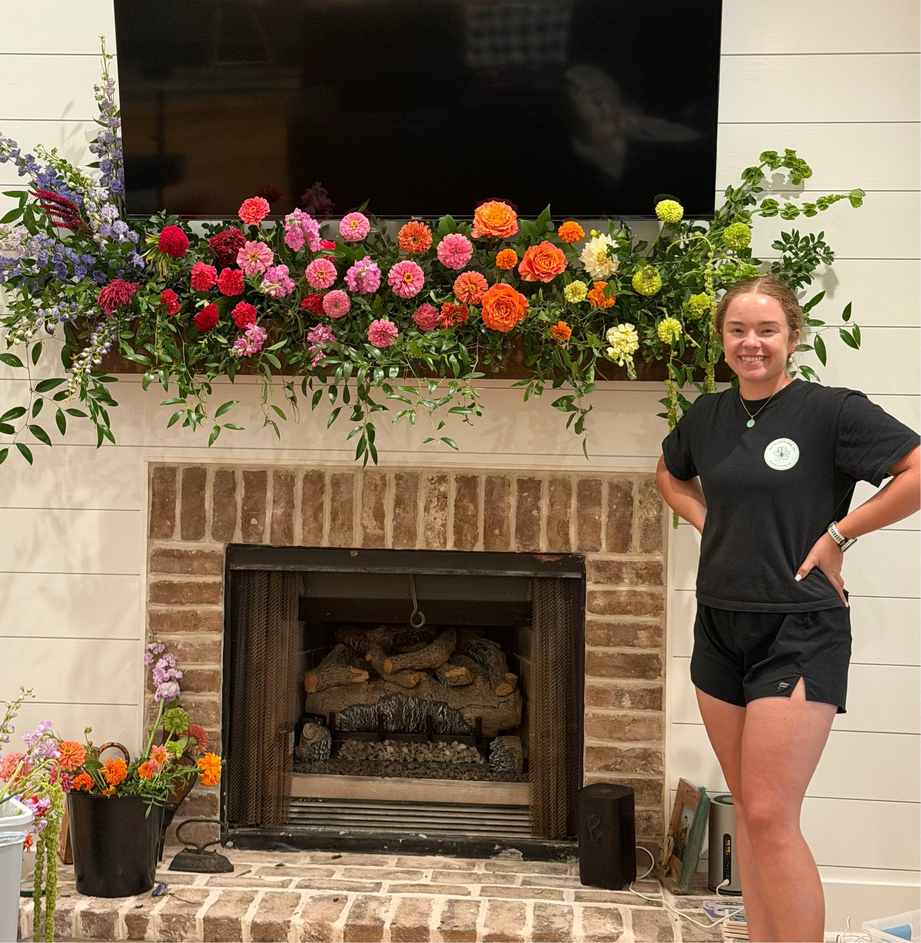 A woman stands to the right of a brick fireplace in a home setting, wearing a black t-shirt and black shorts. The fireplace mantel is decorated with a large, vibrant floral arrangement featuring pink, red, orange, yellow, and purple flowers with green foliage.