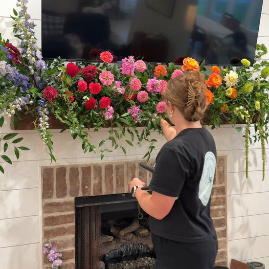 A woman in a black t-shirt and shorts stands in front of a brick fireplace decorated with a large, vibrant floral garland under a mounted television.