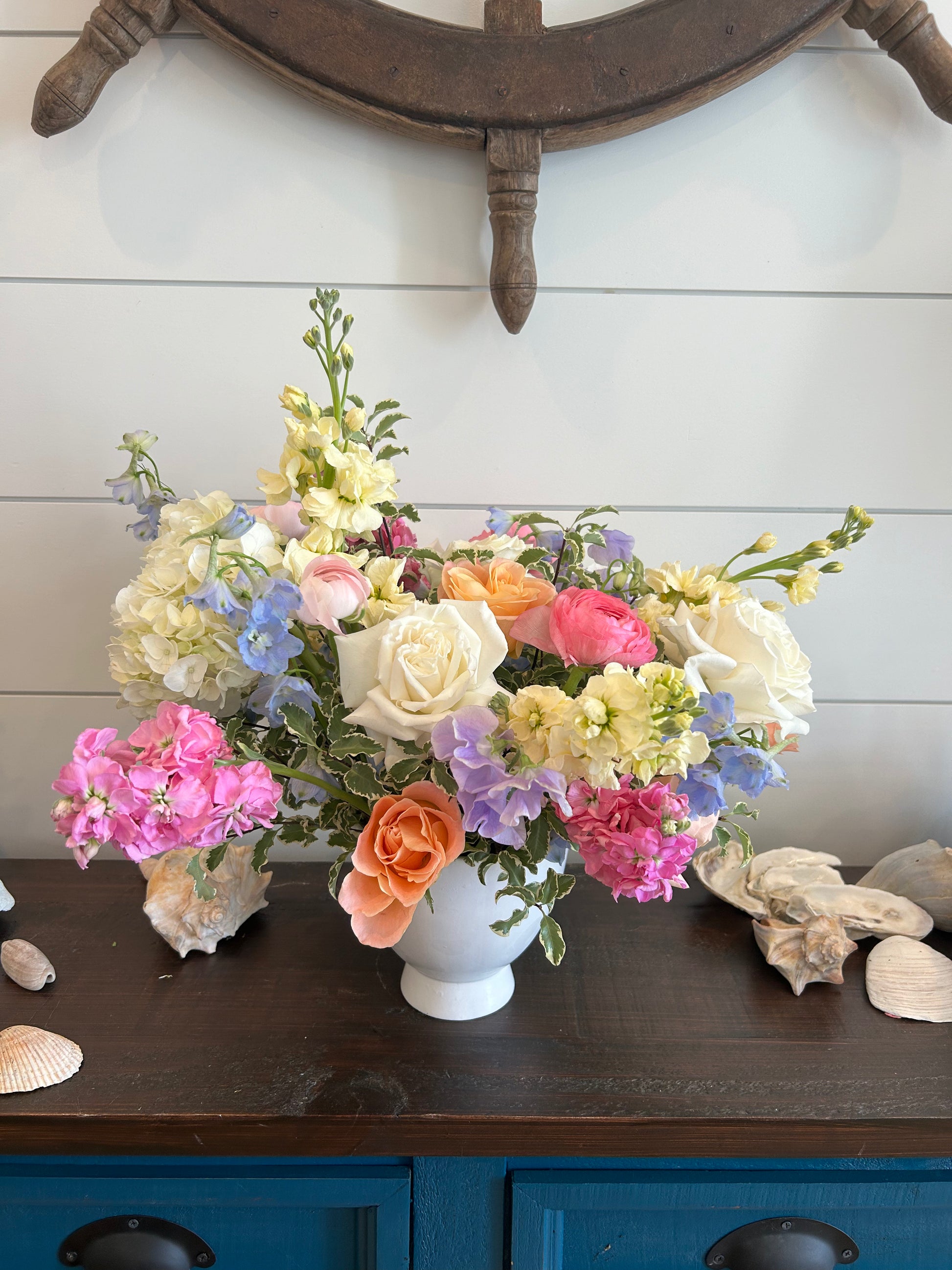 A flower arrangement using pastel colors like light pink, yellow, purple, blue, and white flowers in a white vase.